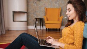 Young adult female sitting on floor against couch shopping online with laptop and debit card